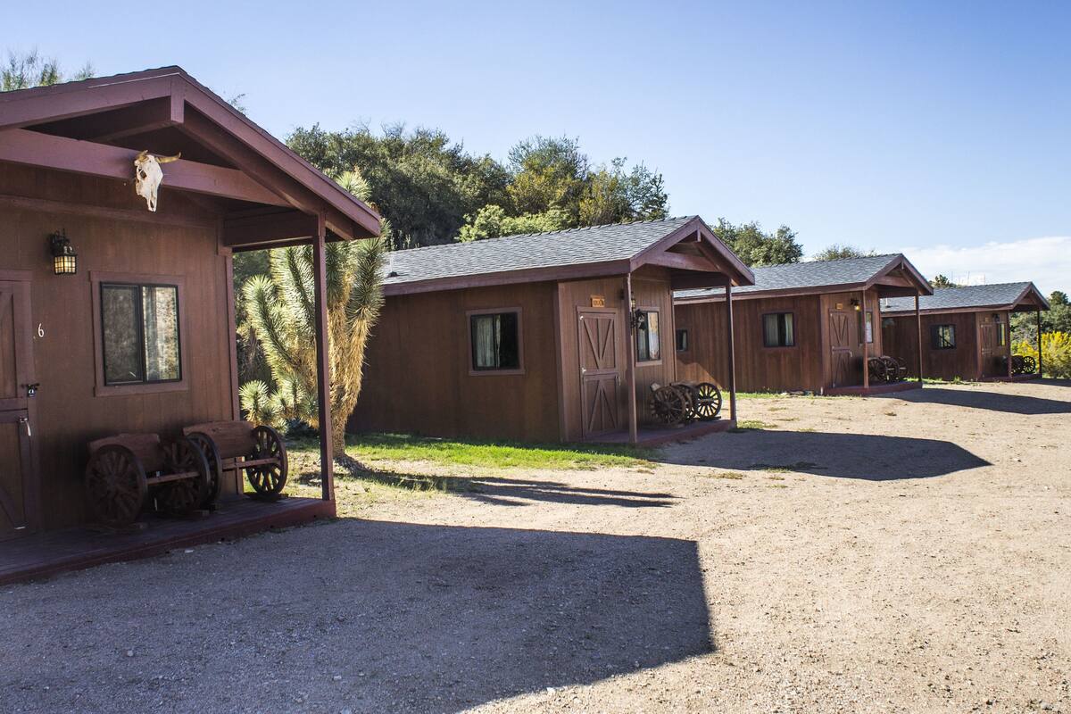 A row of rustic wooden cabins with a gravel driveway, each featuring a door and windows, and a sign indicating the number of rooms.