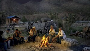 Um grupo de pessoas sentadas ao redor de uma fogueira em um ambiente ao ar livre rústico, com uma estrutura de madeira e um carrinho ao fundo.