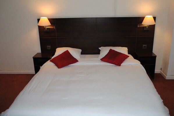Una cama bien tendida con sábanas blancas y dos almohadas rojas, flanqueada por mesitas de noche de madera oscura con lámparas de pared.