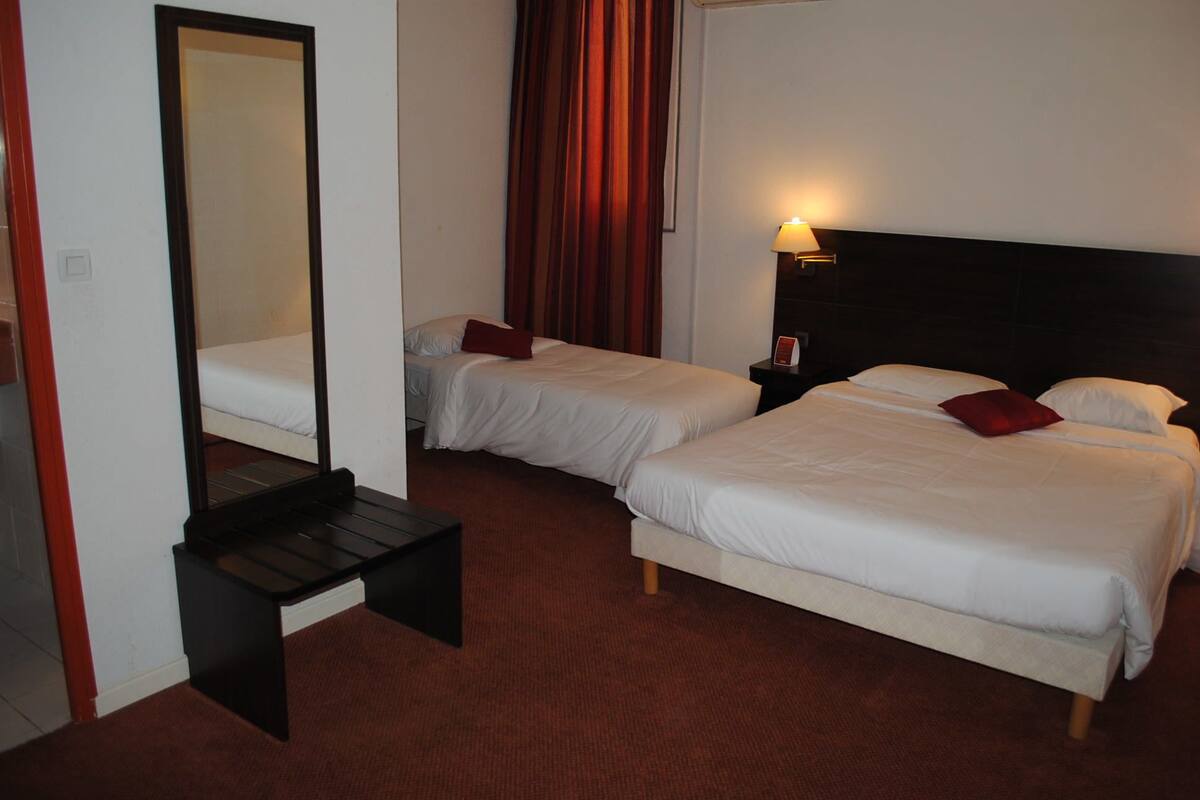 A hotel room with two beds, a desk, a mirror, a nightstand with a lamp, and a red curtain.