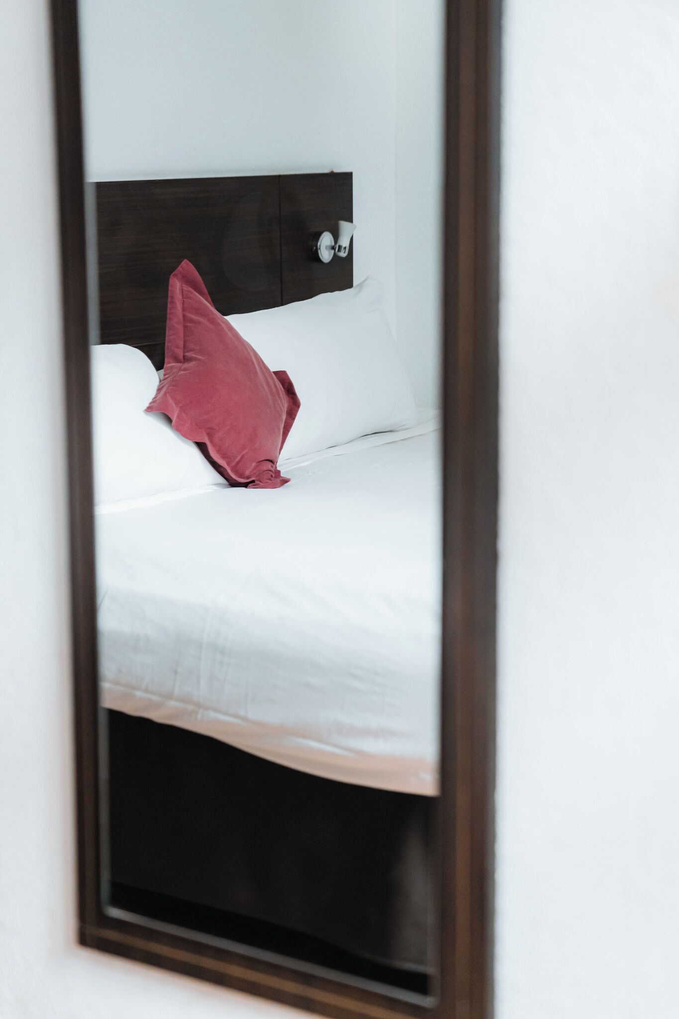 Cama com roupa de cama branca e um único travesseiro vermelho, emoldurada por uma cabeceira de madeira escura.