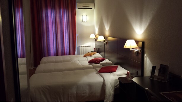 A hotel room with two beds, red and white bedding, a nightstand with a lamp, and a window with purple curtains. - urban by balladins Perpignan (Perpignan)