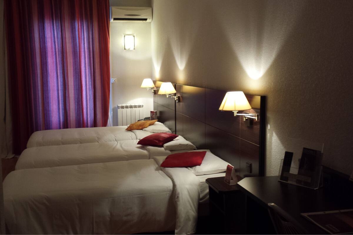 A hotel room with two beds, red and white bedding, a nightstand with a lamp, and a window with purple curtains.