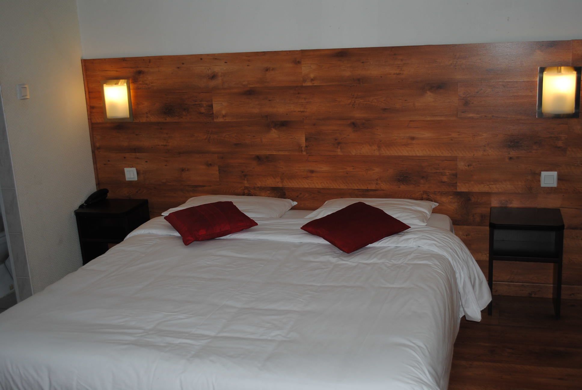 Habitación de hotel con cabecera de madera, dos mesitas de noche, cama con ropa de cama blanca y almohadas rojas, y luces fijadas a la pared.