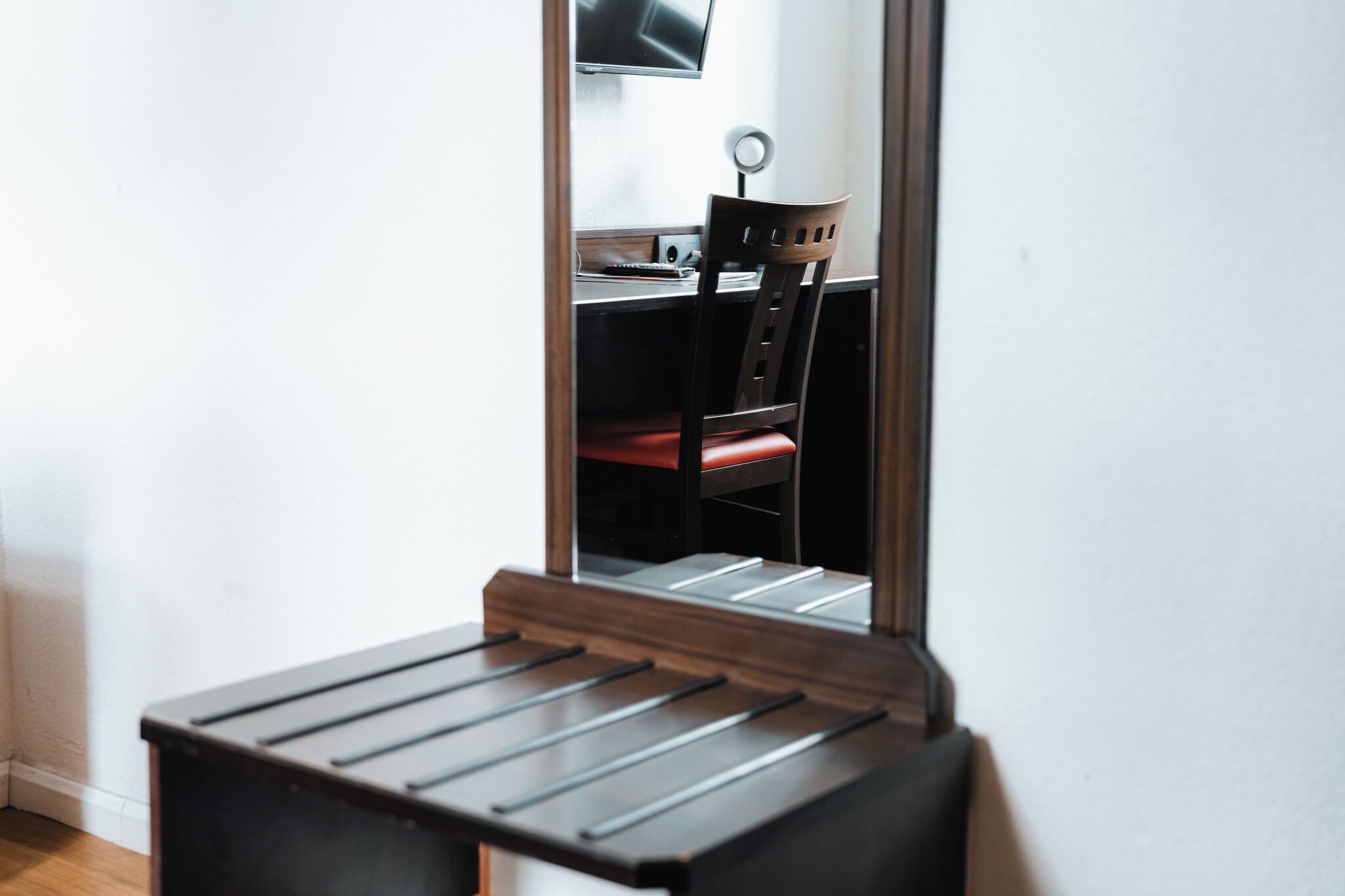 A room with a wooden desk, a chair, and a mirror reflecting the room.