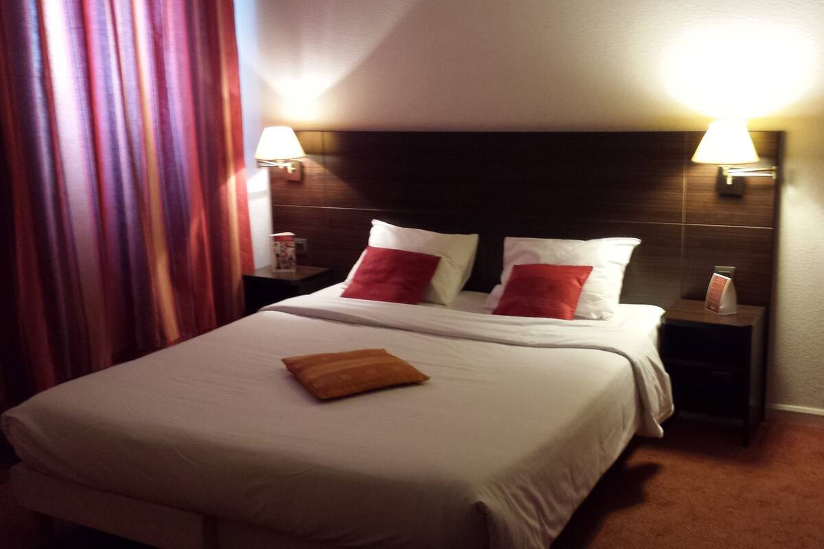 A hotel room with a bed, two bedside tables, a nightstand, a lamp, and a curtain.
