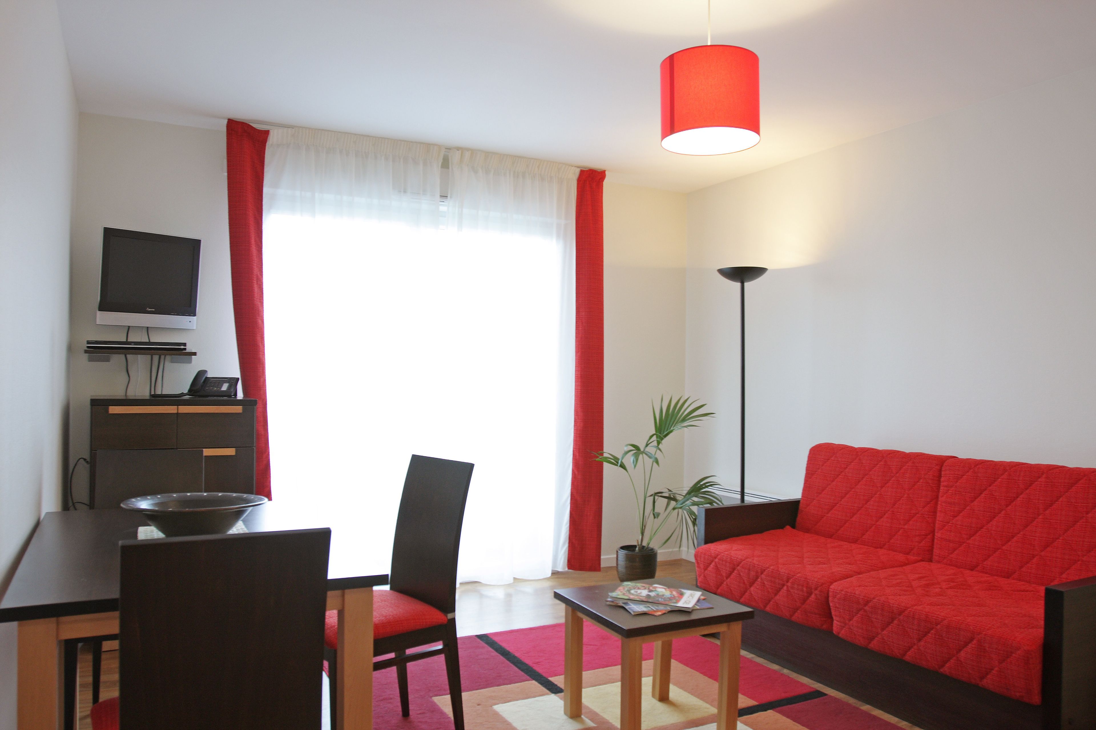 A modern living room with a red sofa, a dining table with chairs, a TV, and a floor lamp.
