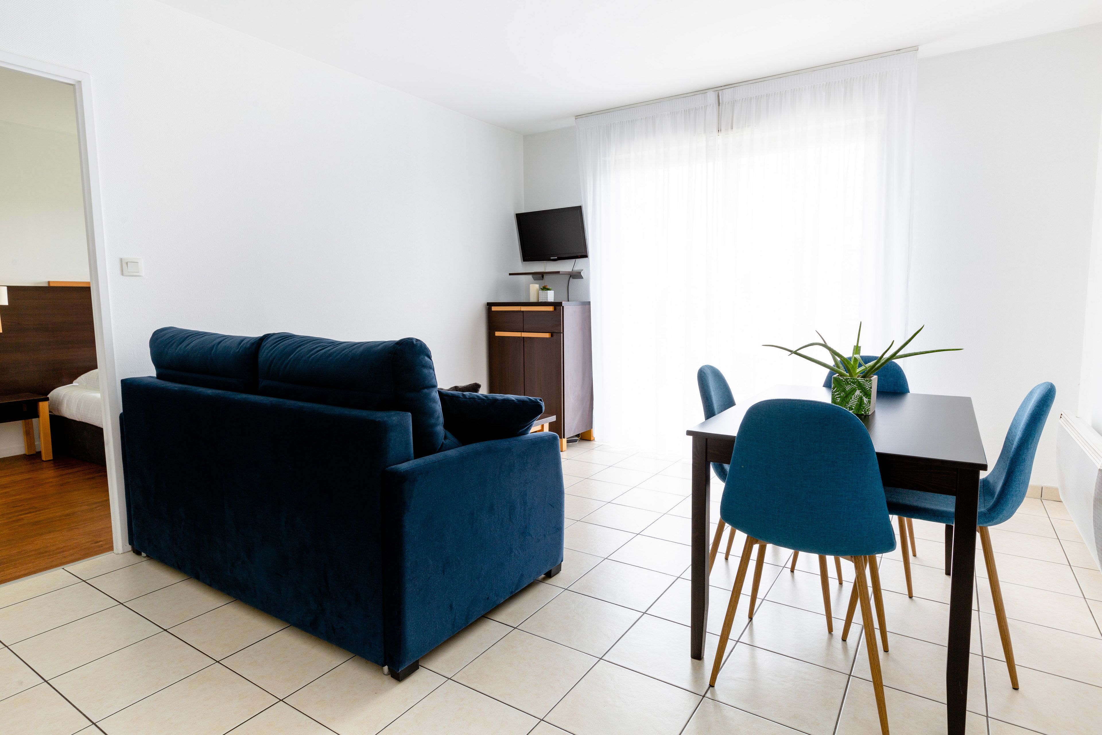A modern living room with a dark blue sofa, a dining table with blue chairs, and a flat-screen TV on a wooden cabinet.