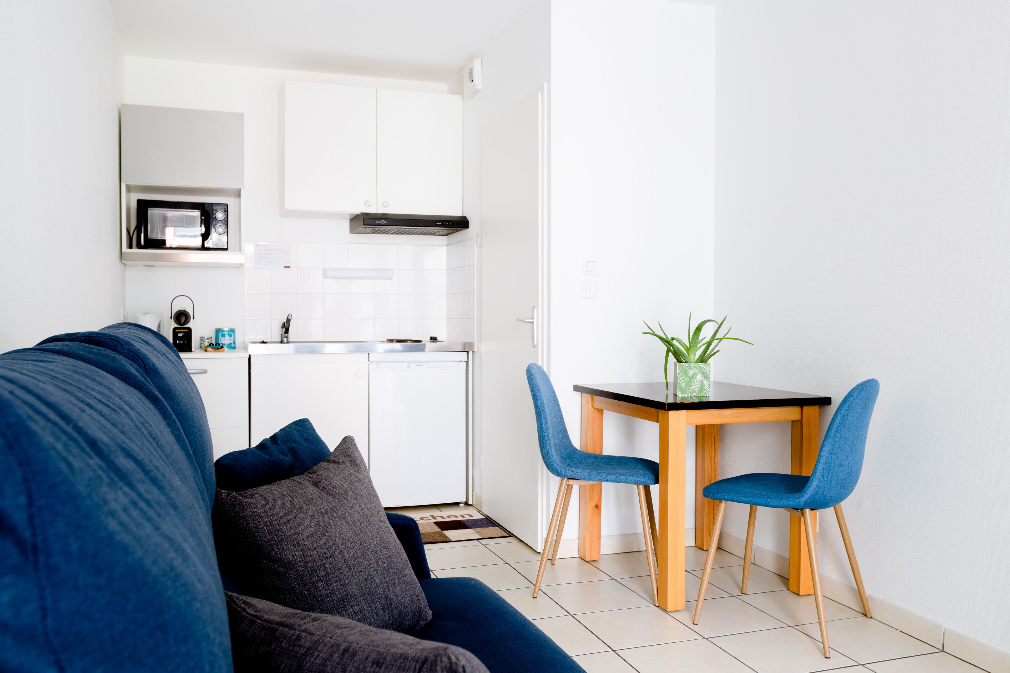 Une cuisine moderne avec des armoires blanches, un four à micro-ondes et un évier. On y trouve un coin repas avec une table en bois et deux chaises bleues. Un canapé bleu foncé se trouve au premier plan.