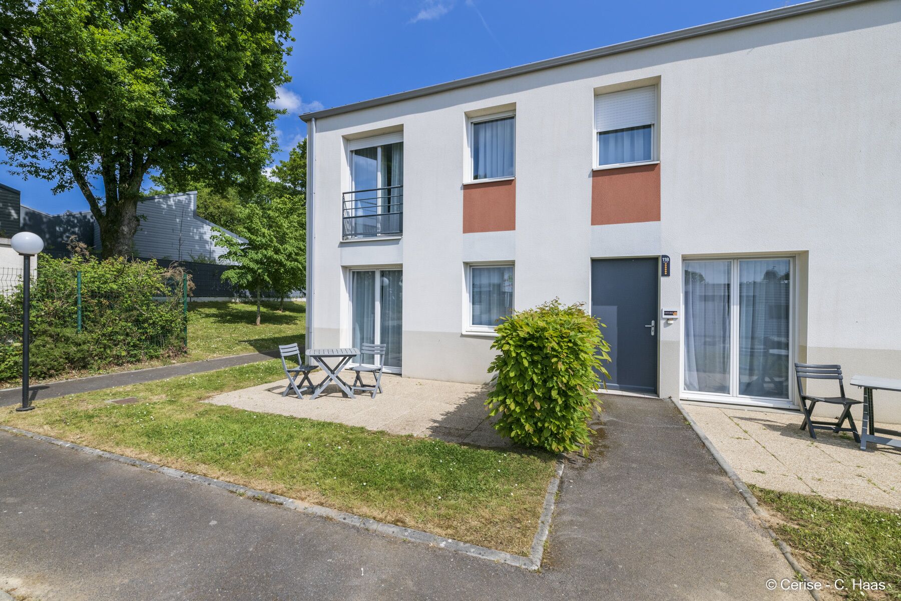 Un edificio de apartamentos de dos pisos con fachada plana, ventanas grandes y un pequeño patio con mobiliario exterior.