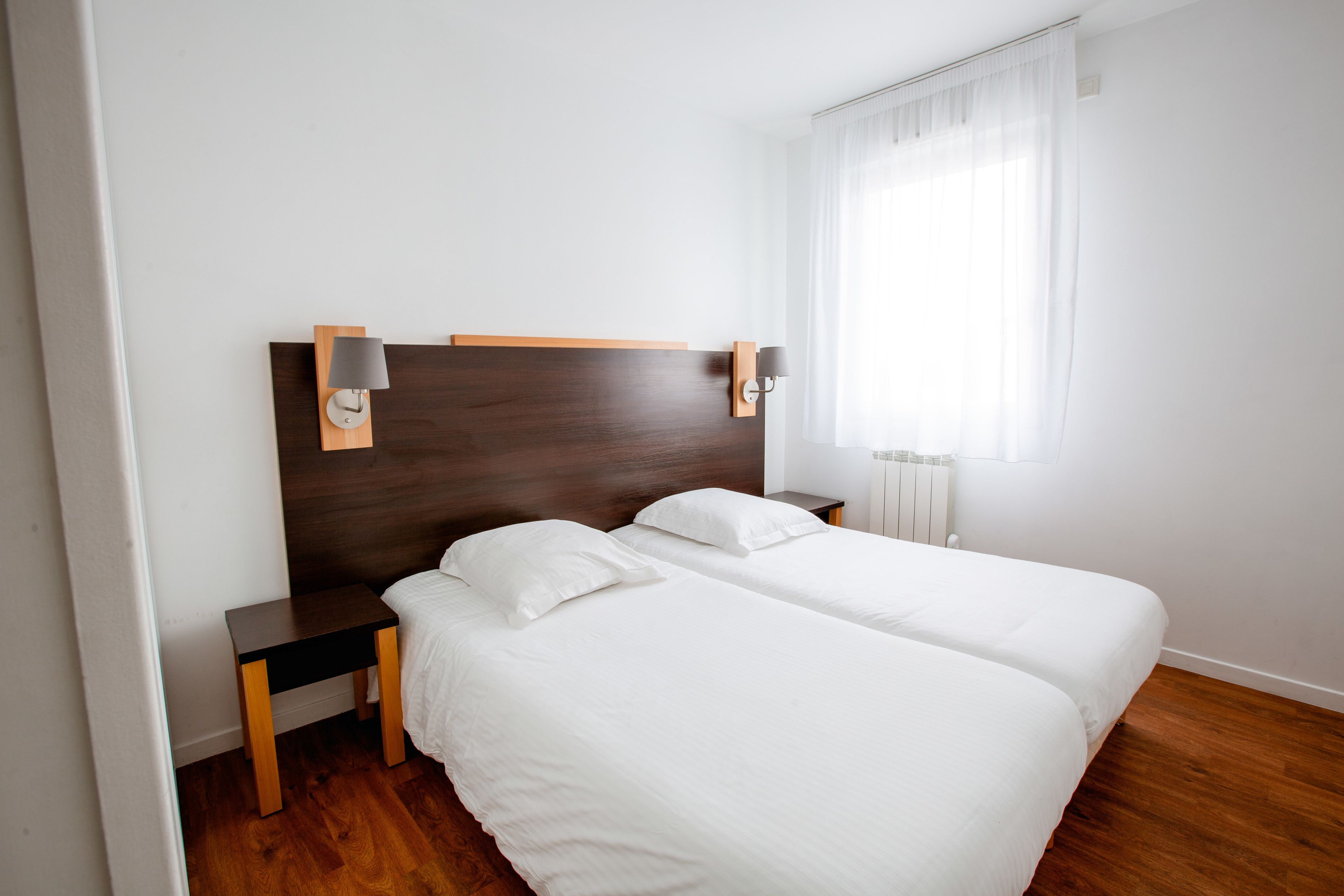 Ein Hotelzimmer mit zwei Betten, einem Holzkopfteil, Nachttischen und einem Fenster mit durchscheinenden Vorhängen.