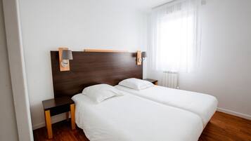 Ein Hotelzimmer mit zwei Betten, einem Holzkopfteil, Nachttischen und einem Fenster mit durchscheinenden Vorhängen.