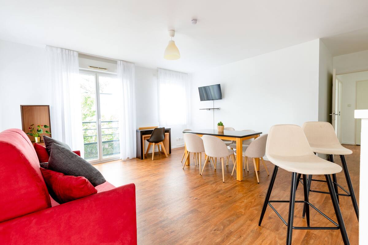 Una sala de estar moderna con un sofá rojo, una mesa de comedor con sillas, un televisor montado en la pared y un balcón visible a través de la ventana.