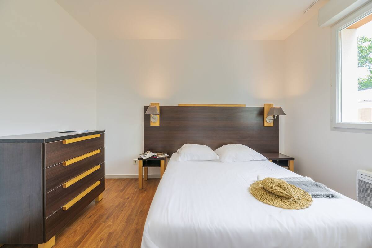 Un dormitorio con un aparador de madera, una cama con sábanas blancas, un sombrero de paja sobre la cama y una mesita de noche con una lámpara.