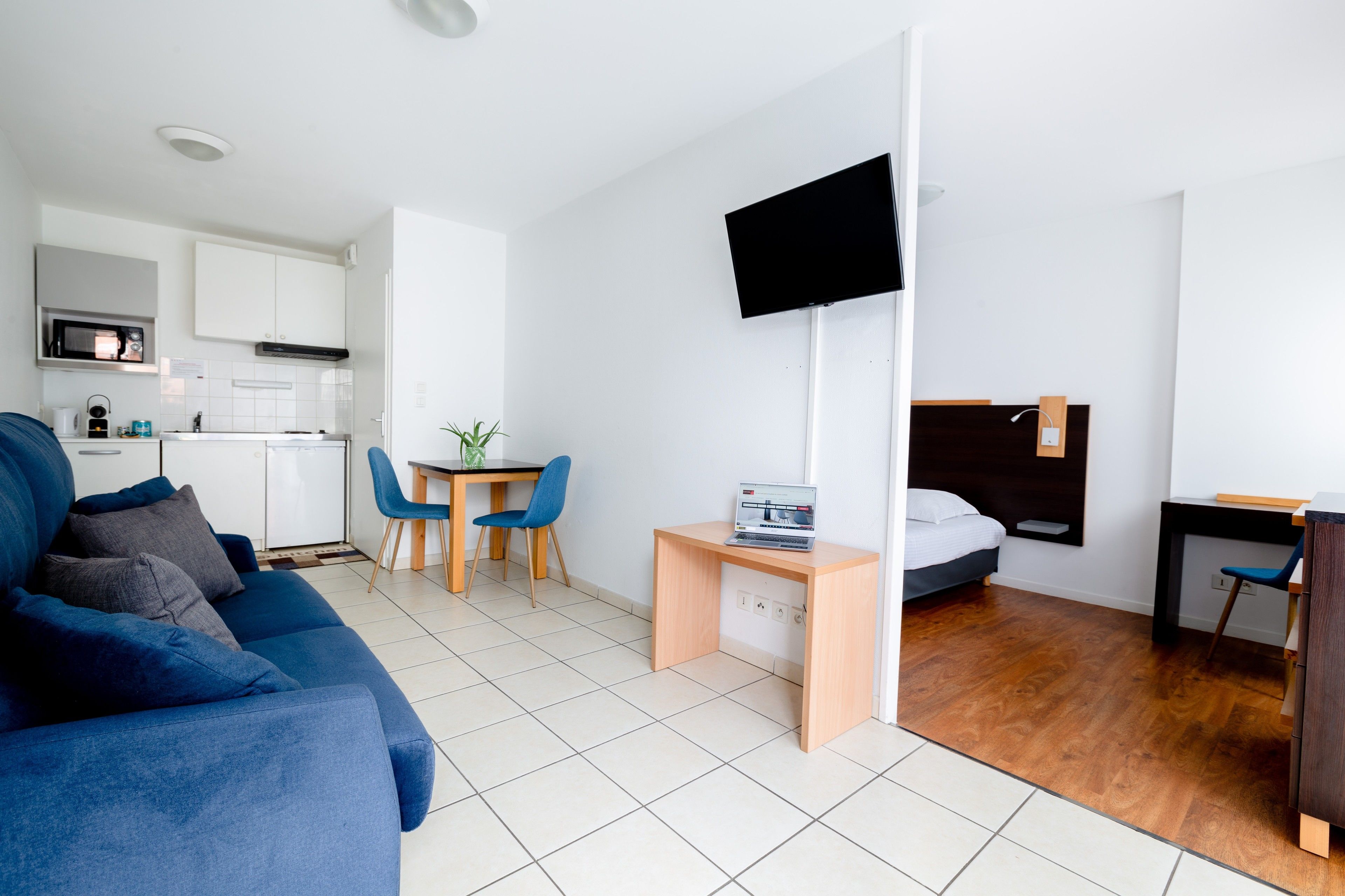 A modern, well-lit room with a blue sofa, a small desk with a laptop, a TV mounted on the wall, and a bed with a wooden headboard.