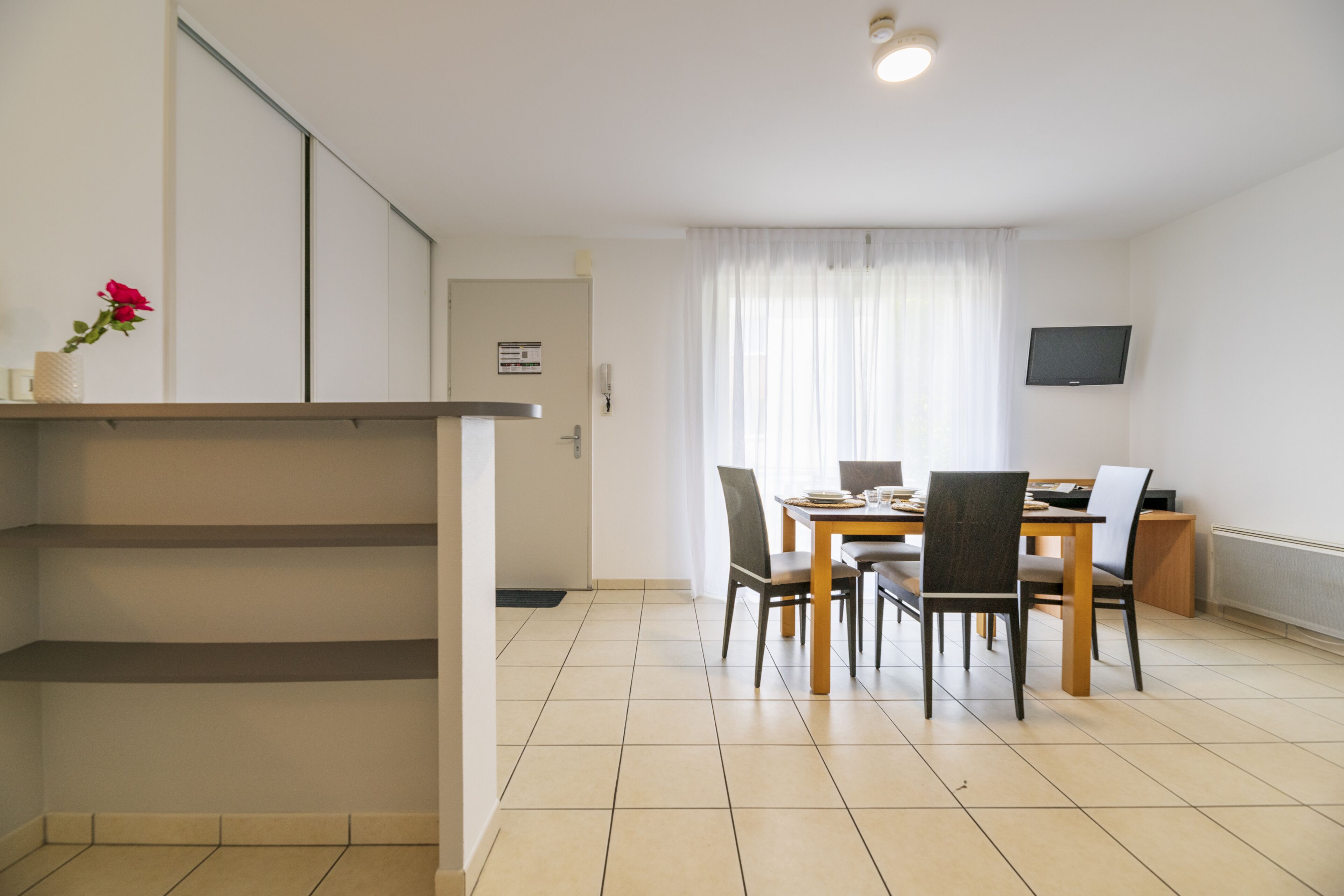 A modern kitchen with a dining area, a TV mounted on the wall, and a window with sheer curtains.
