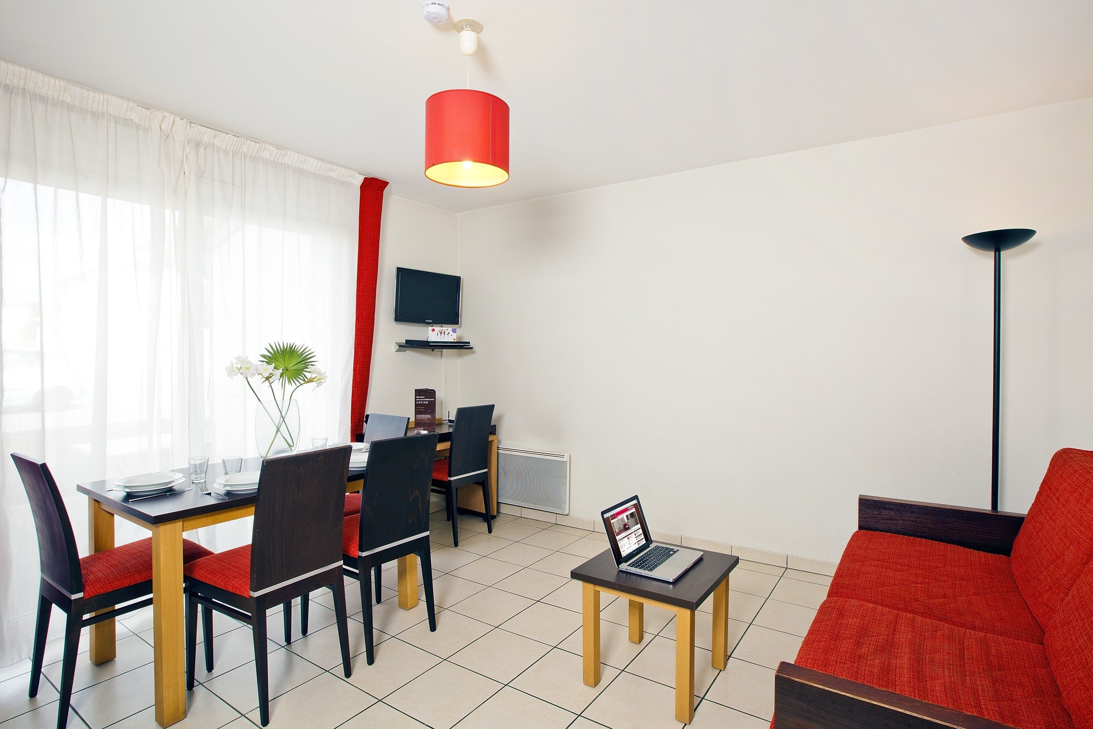 Una sala de estar moderna con un sofá rojo, una mesa de comedor con sillas, una pequeña mesa de centro con una computadora portátil, y un televisor en la pared.