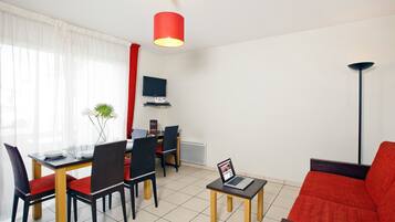 Una sala de estar moderna con un sofá rojo, una mesa de comedor con sillas, una pequeña mesa de centro con una computadora portátil, y un televisor en la pared.