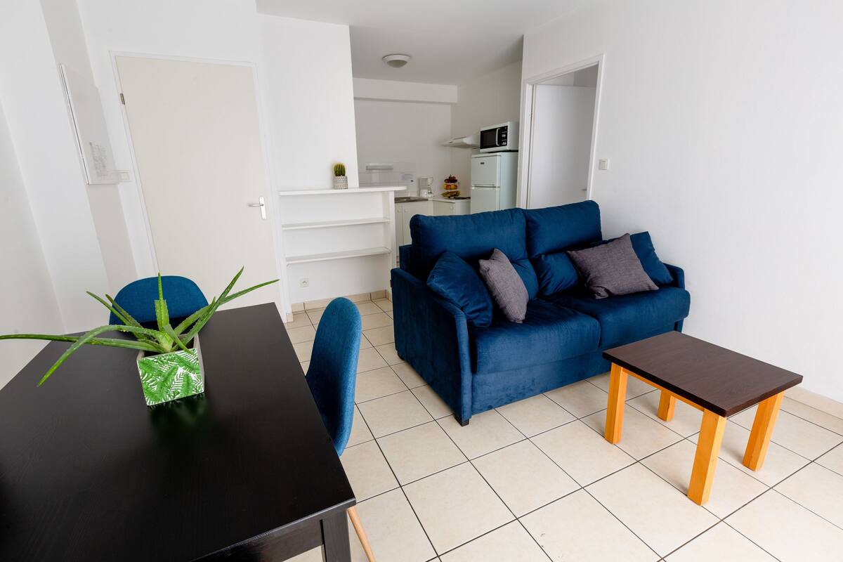 Un moderno salón con un sofá azul, una pequeña mesa de madera y una mesa de centro negra. En el fondo, se ve una cocina blanca.