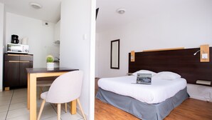 Una habitación de hotel moderna con suelo de madera, cama con falda gris, escritorio con silla, una pequeña planta en maceta y una kitchenette con microondas y cafetera.
