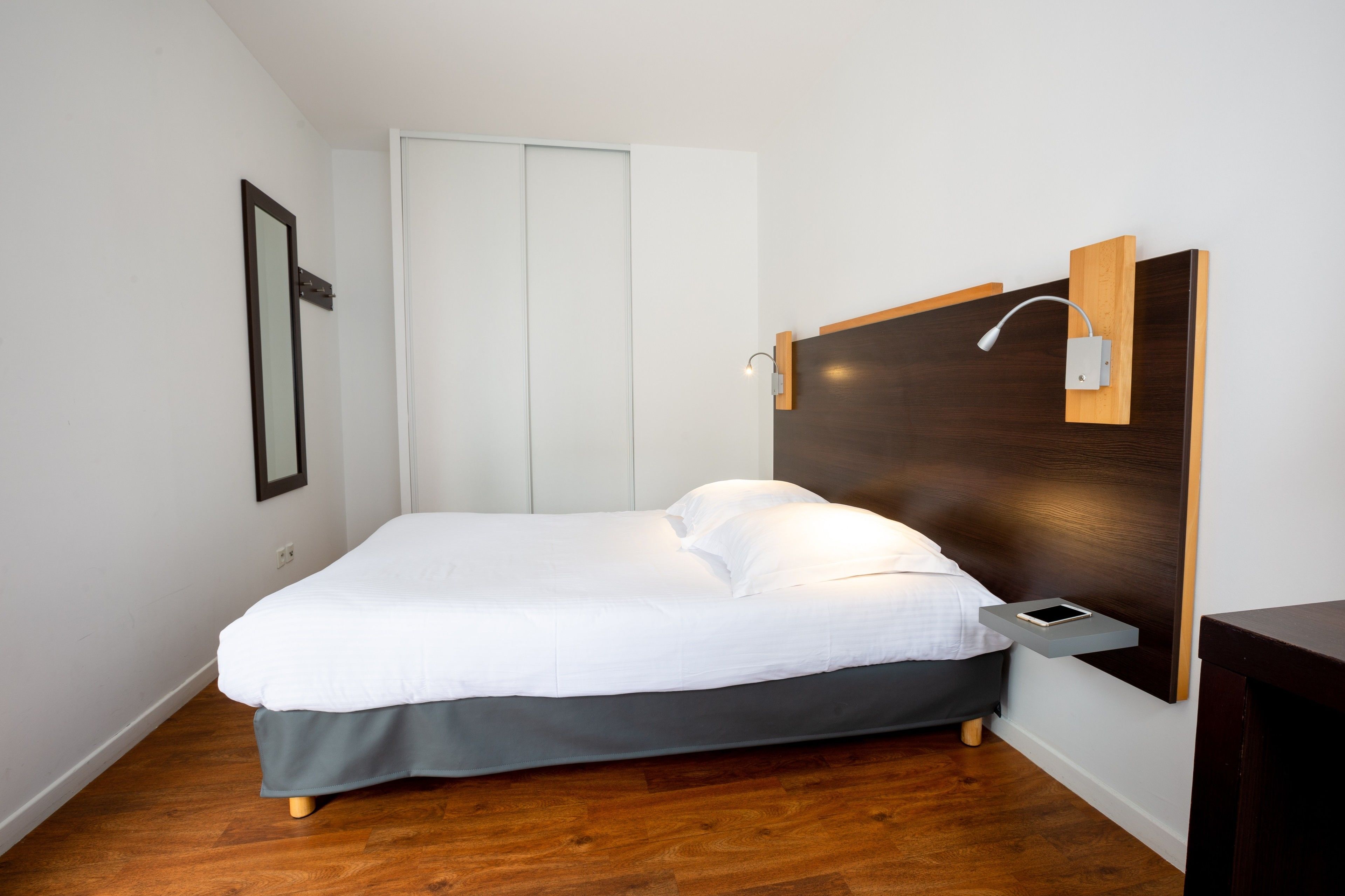 A neatly made bed in a room with a wooden floor, a bedside table with a lamp, and a wall-mounted shelf.