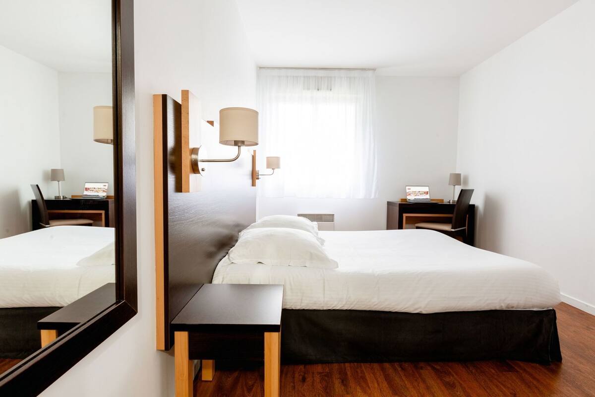 Una habitación de hotel con una cama grande, un escritorio, una silla, un espejo y una ventana con cortinas.