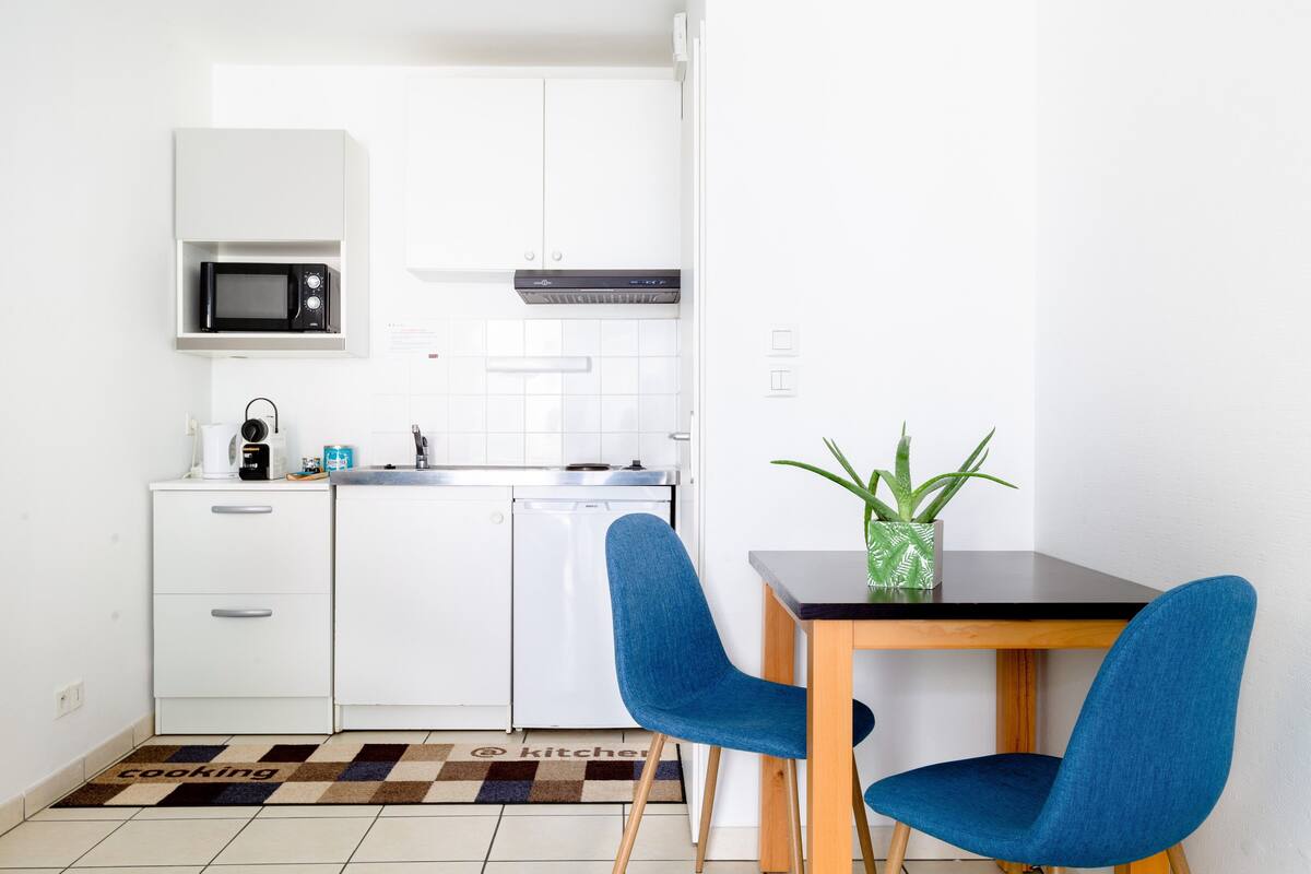 Una cocina compacta con armarios blancos, un microondas, una cafetera y un lavavajillas. Hay una mesa de comedor con dos sillas azules y una planta en maceta.