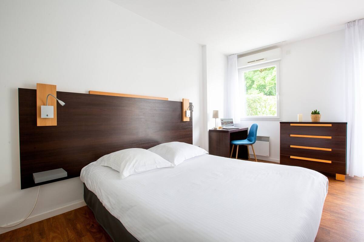 Una habitación de hotel moderna con una cama grande, un escritorio, una silla, un aparador y una ventana con vista a zonas verdes.