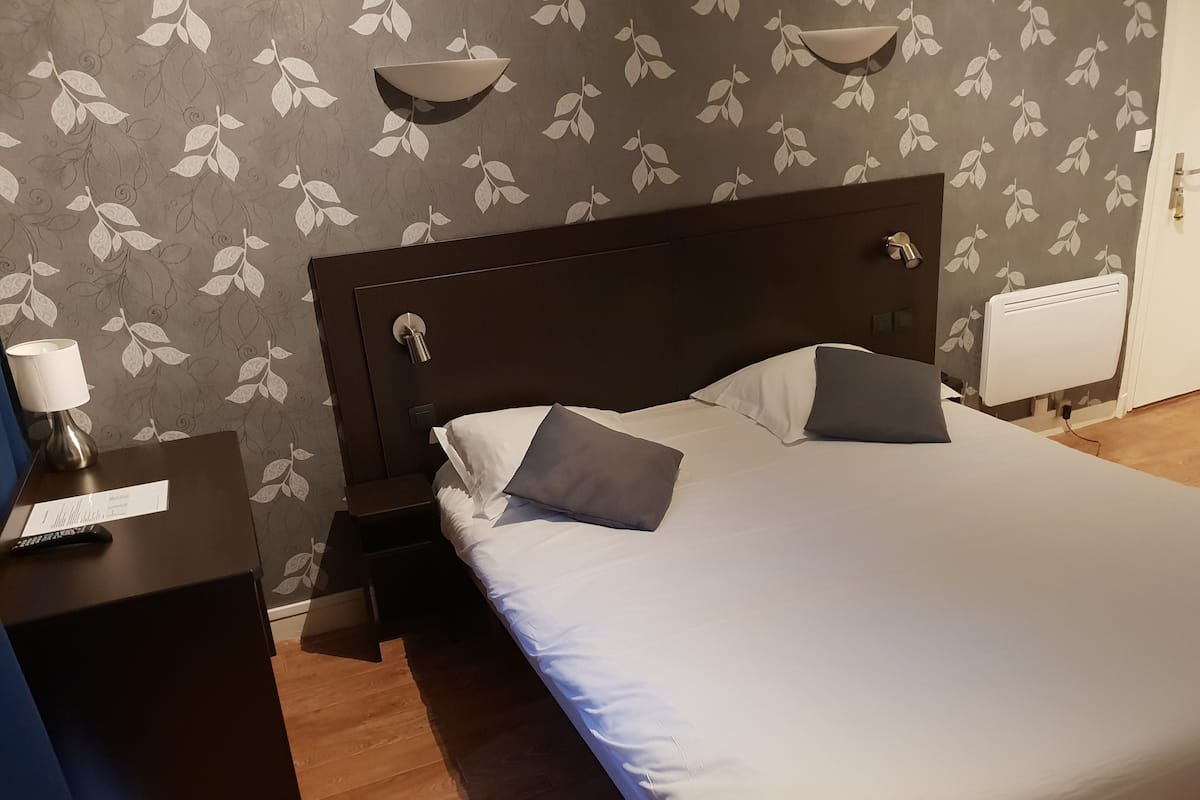 A neatly made bed with white and gray bedding, a dark wooden headboard, and a nightstand with a lamp and remote control.