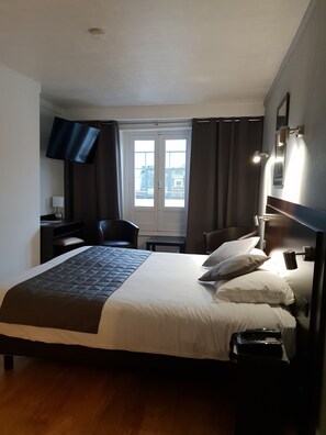 A hotel room with a large bed, a desk, a chair, a TV, and a window with curtains. - Le Napoleon (Lille)