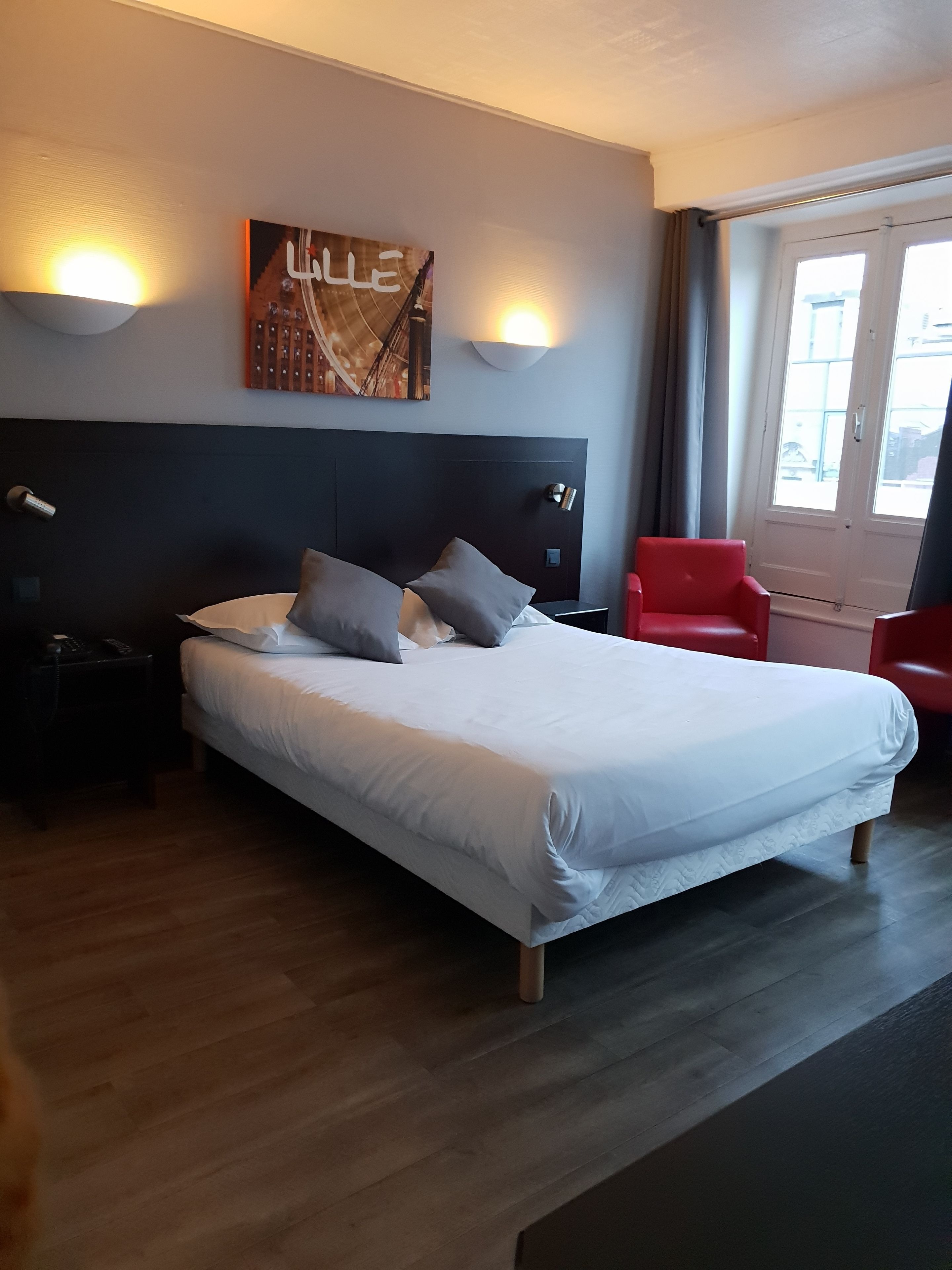 Une chambre d’hôtel avec un grand lit, une tête de lit, deux lampes de chevet, une chaise rouge et une fenêtre avec des rideaux.