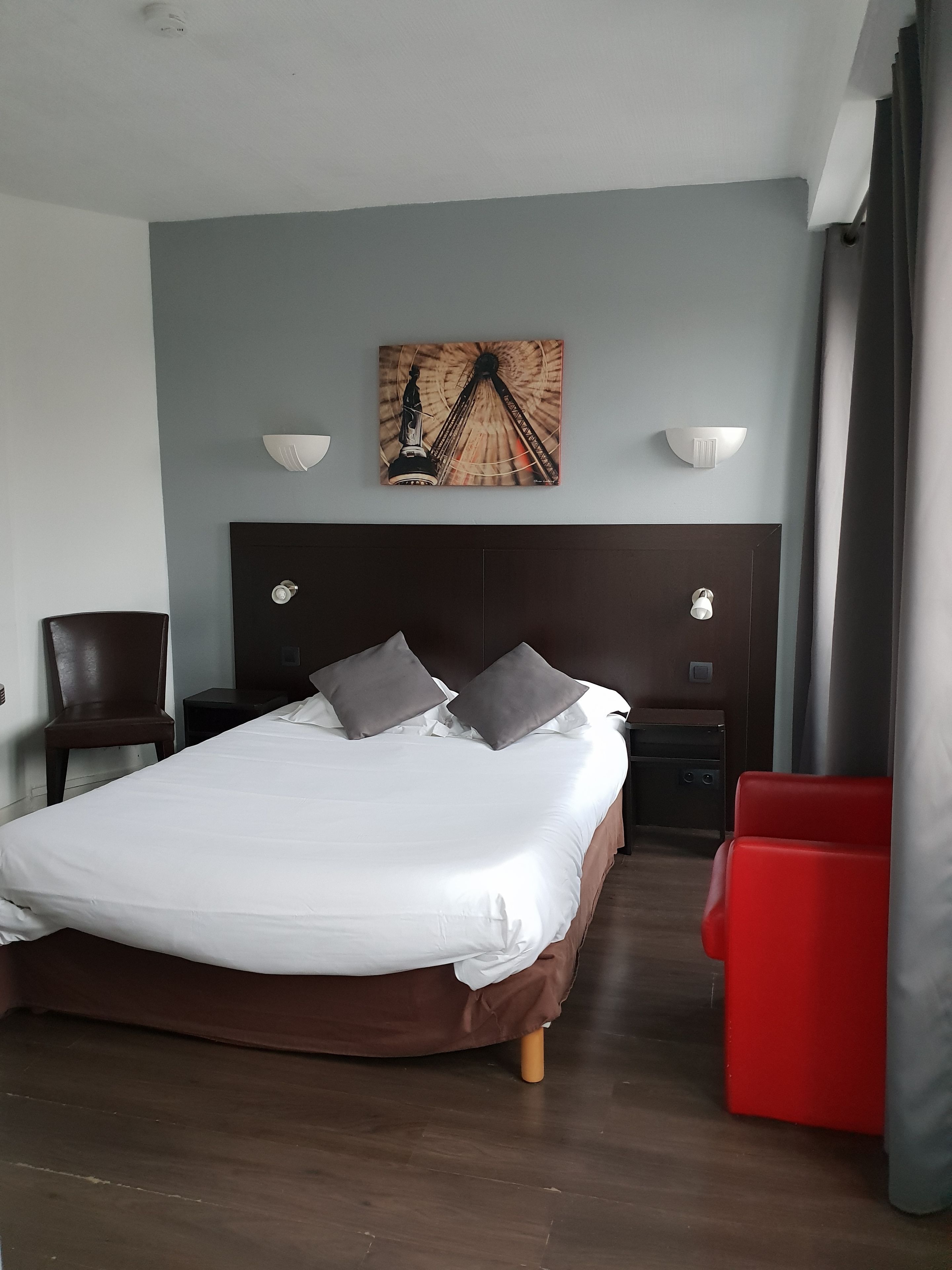 A hotel room with a large bed, two bedside tables, a headboard, a red chair, a wall-mounted artwork, and a window with curtains.