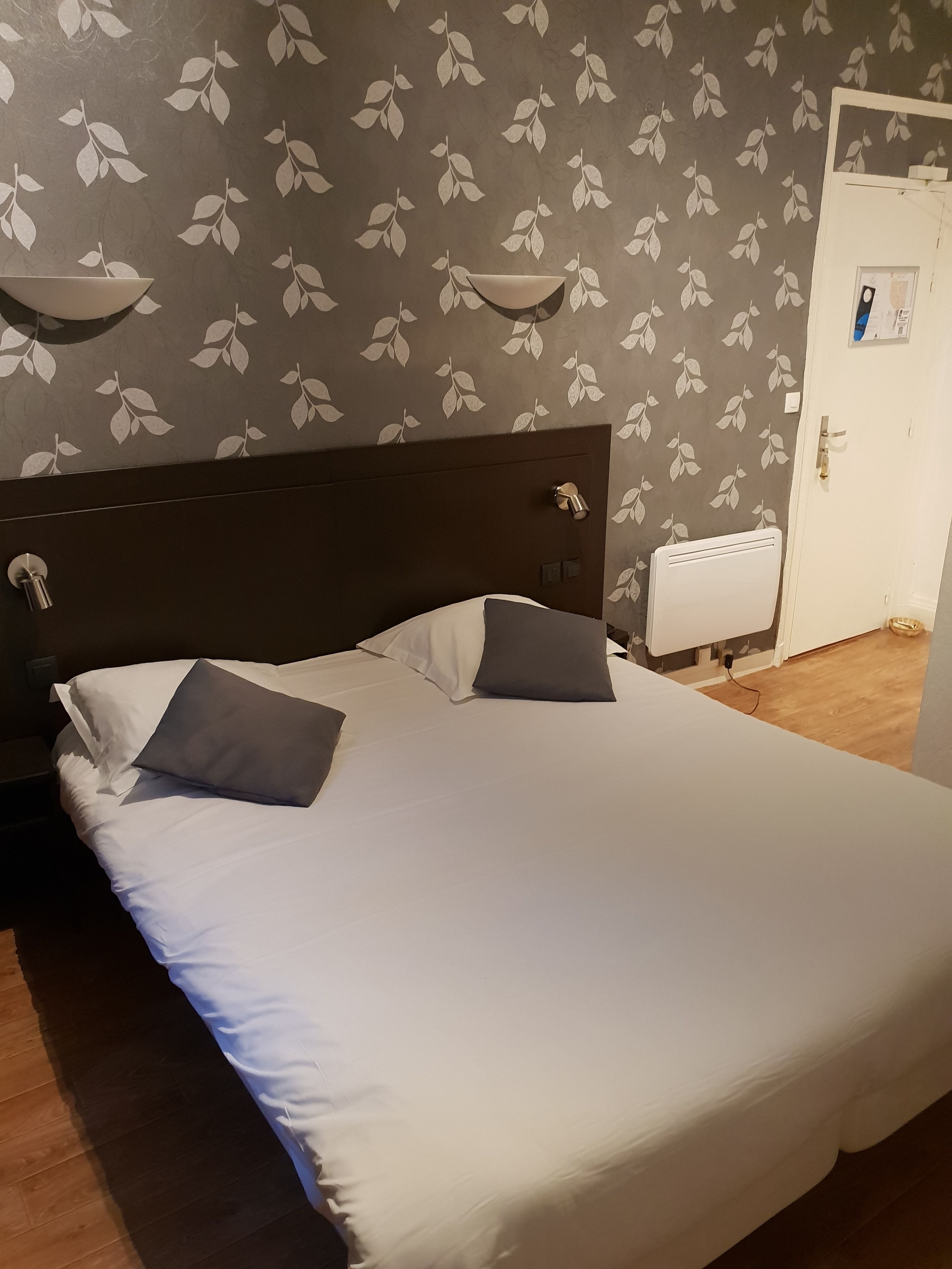 A neatly made bed with white and gray linens, a dark wooden headboard, and a wall with a leaf pattern.