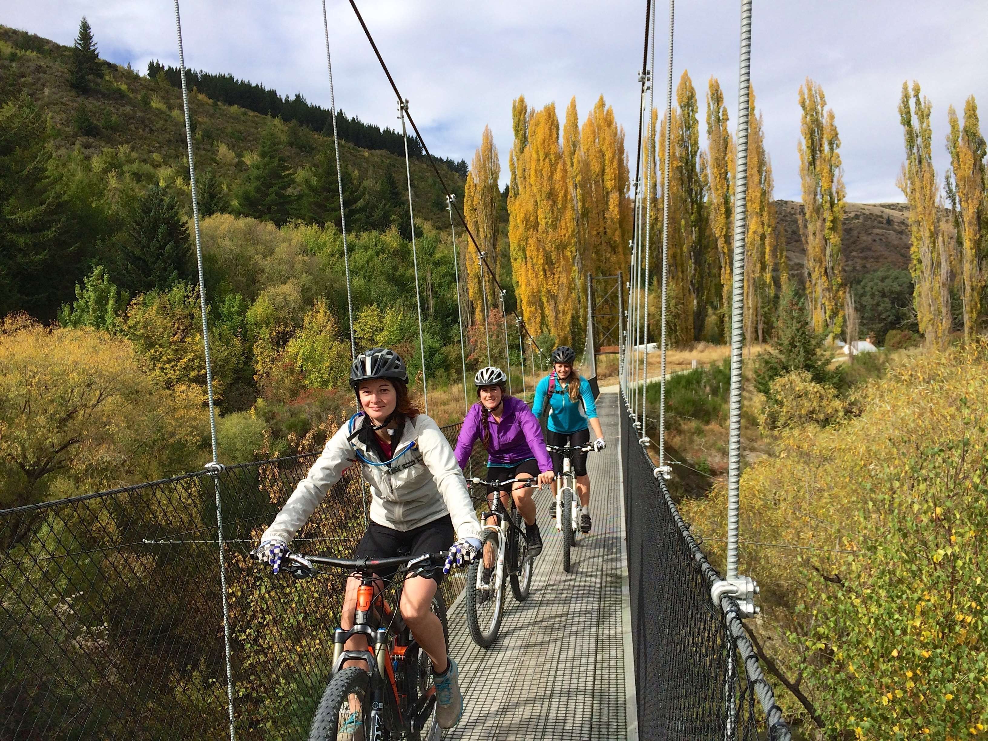 Three cyclists on a suspension bridge surrounded by autumn foliage.