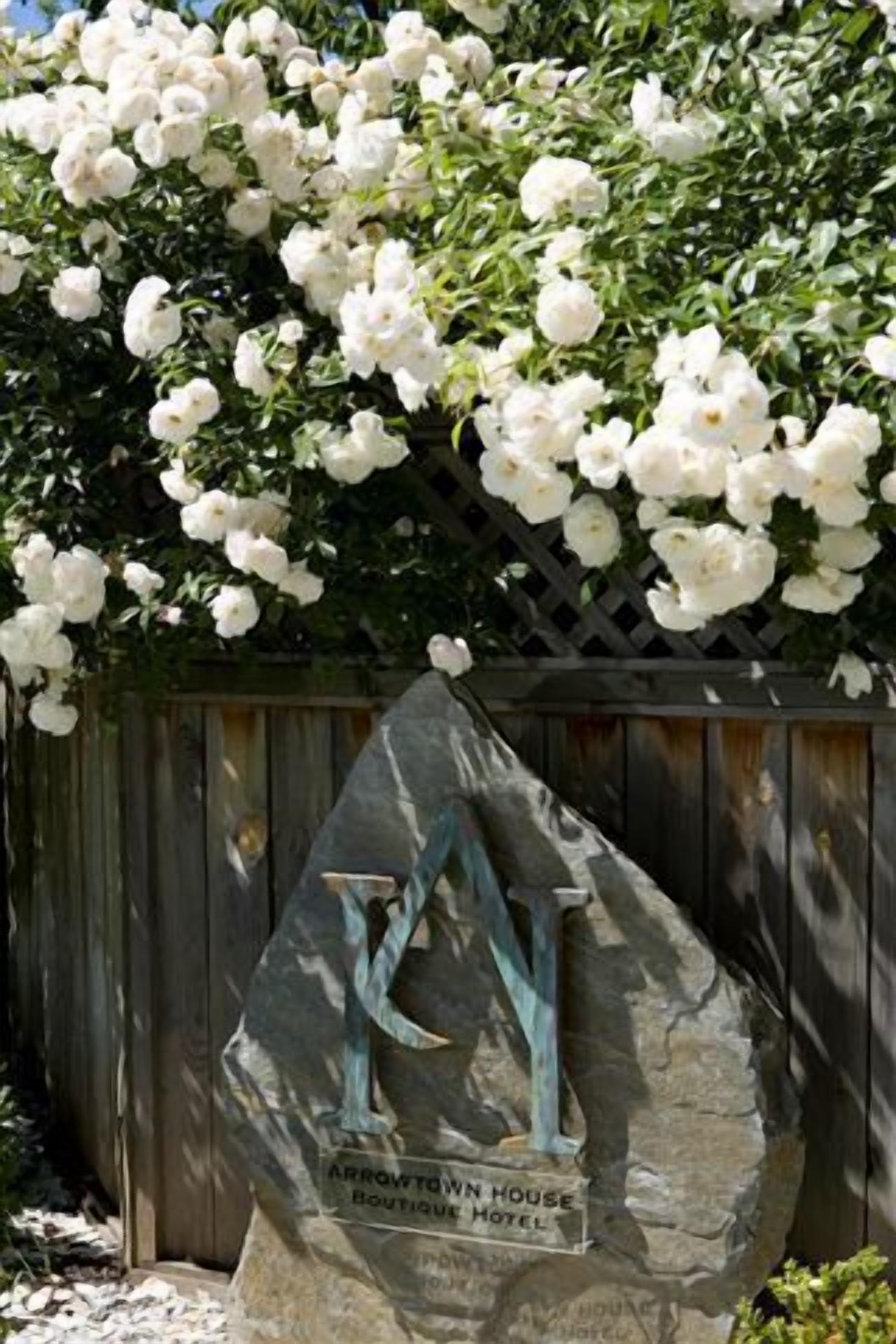 A stone sculpture with a plaque reading "Arrowtown House Boutique Hotel" in front of a wooden fence with white flowering plants.