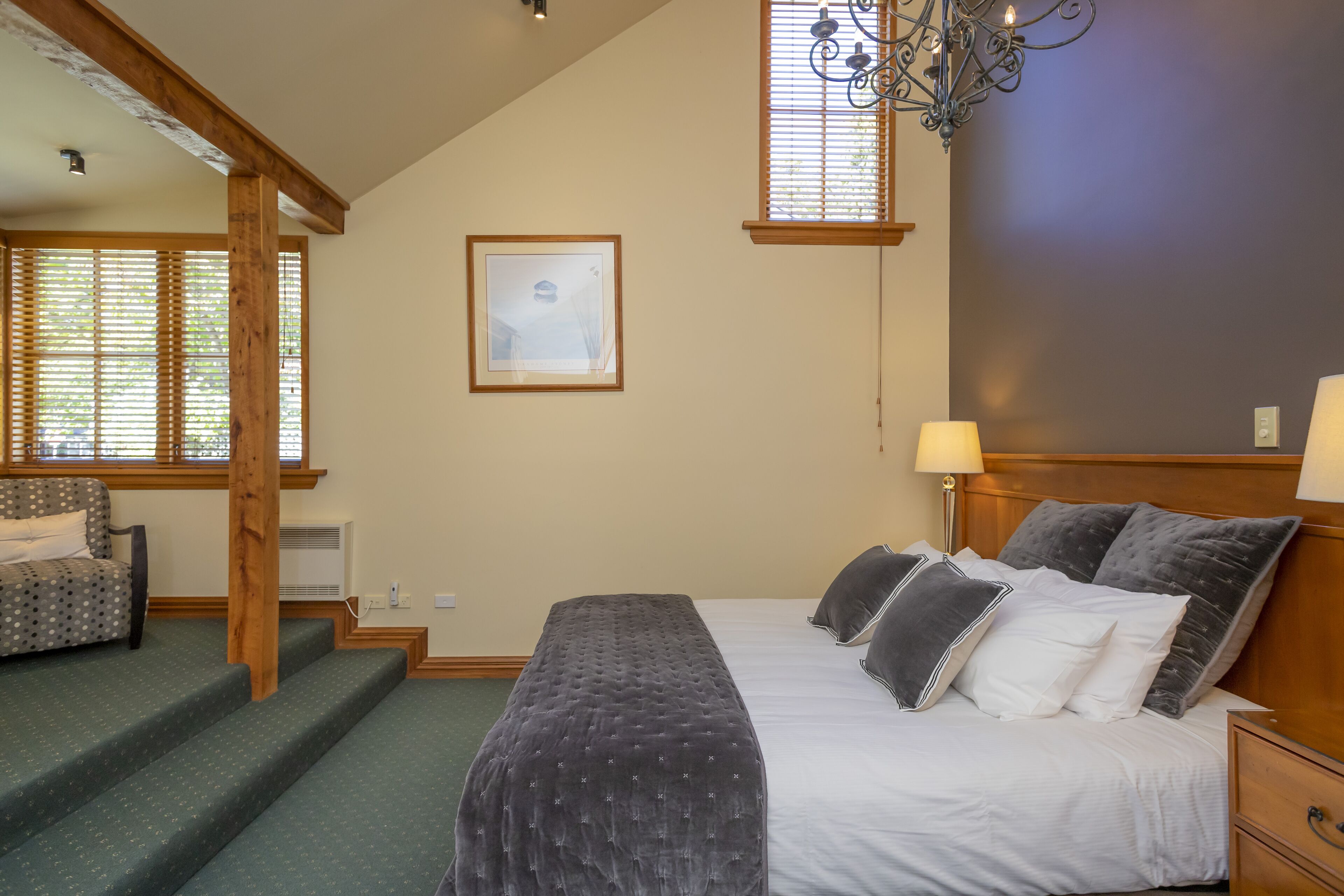 A bedroom with a bed, bedside tables, a chair, a staircase, a chandelier, and a window with blinds.