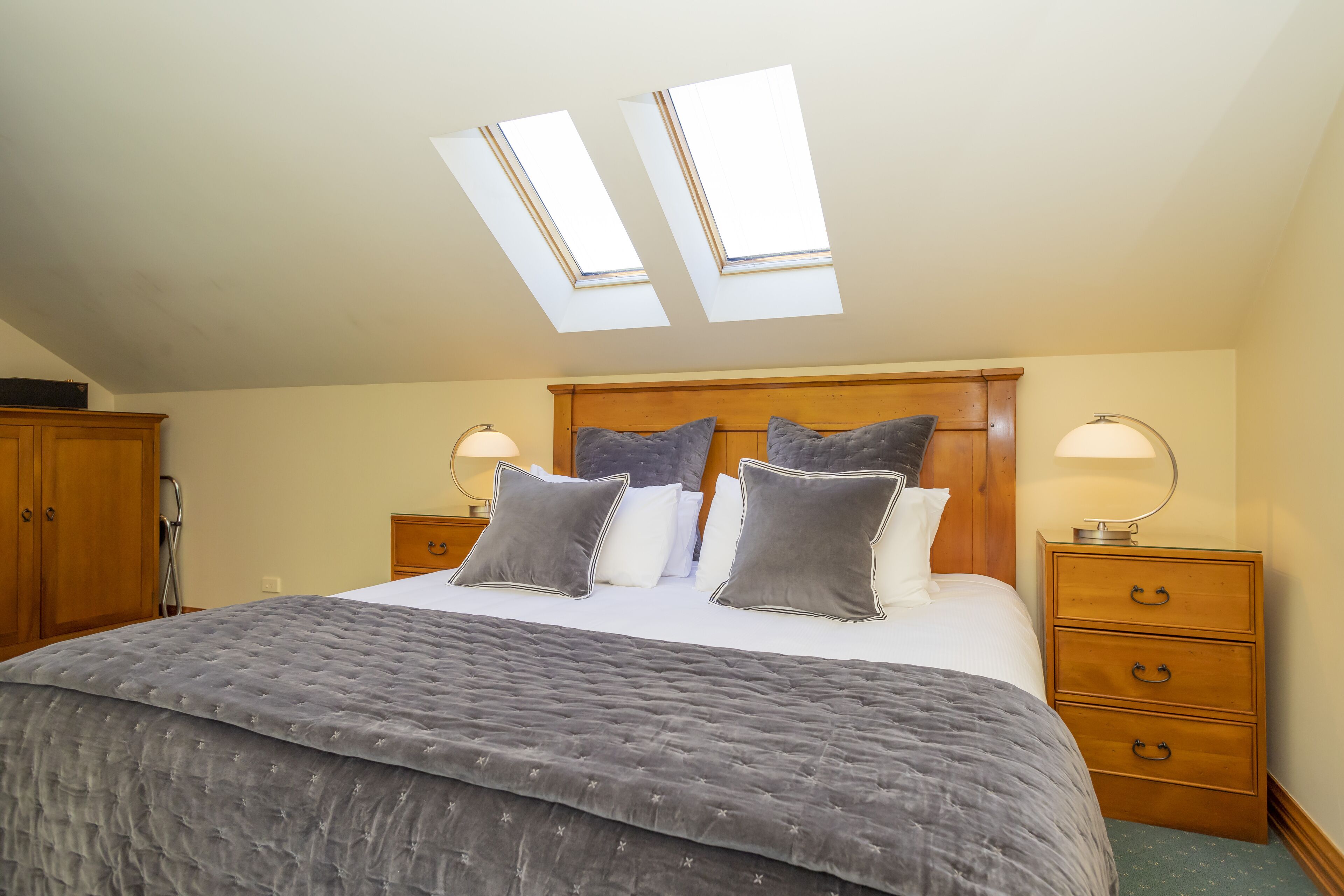 A bedroom with a large bed, skylights, wooden headboard, bedside tables with lamps, and a wooden wardrobe.