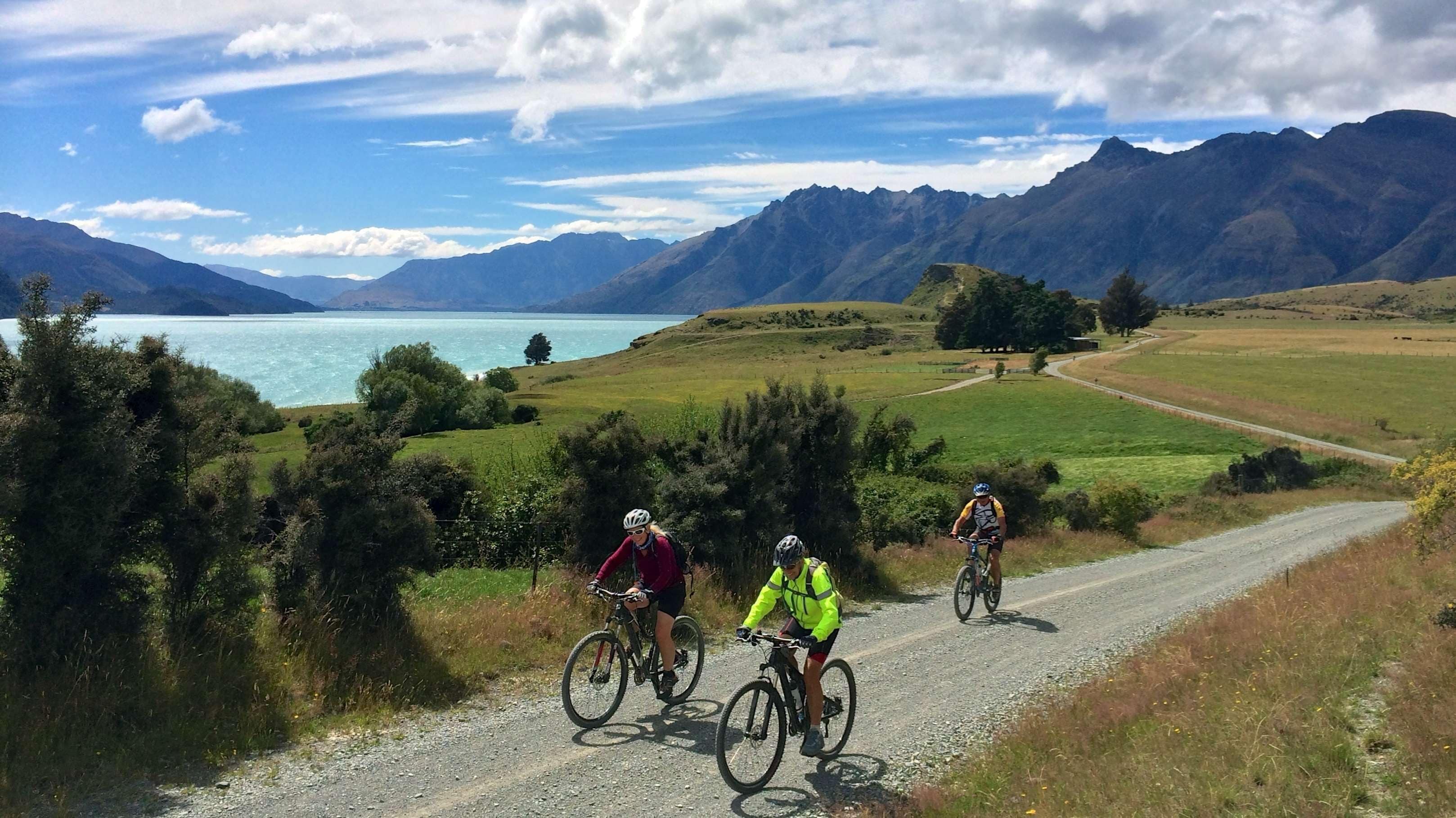 Un chemin de gravier bordé de cyclistes, un lac et des montagnes en arrière-plan.