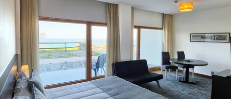 A hotel room with a bed, a sofa, a table, and chairs. There are large windows with curtains, a view of the outdoors, and a ceiling light fixture.