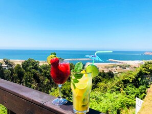 Two drinks on a wooden railing with a scenic view of the sea and greenery in the background.