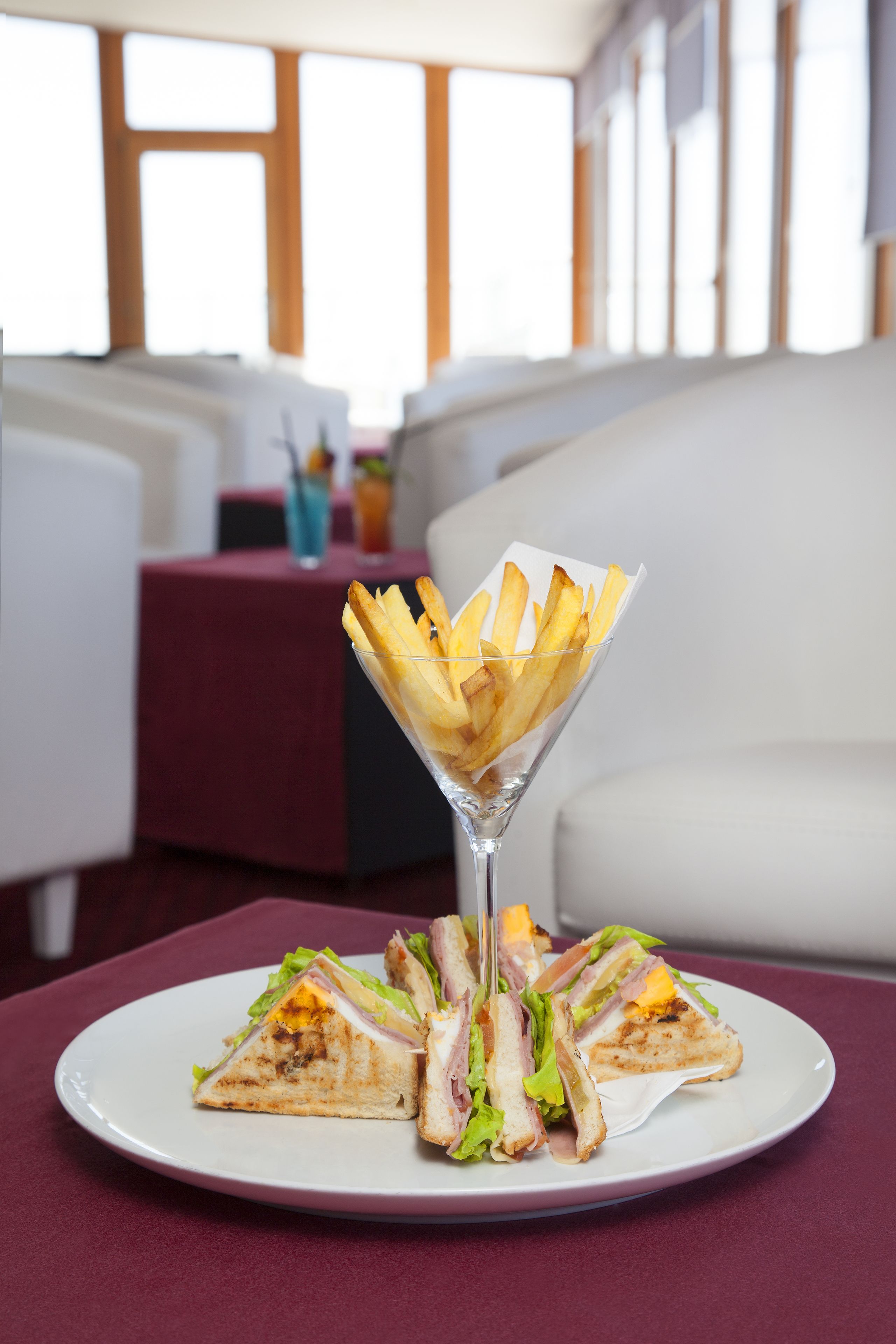 A plate with a sandwich and fries on a table in a dining area with a red tablecloth and white furniture.