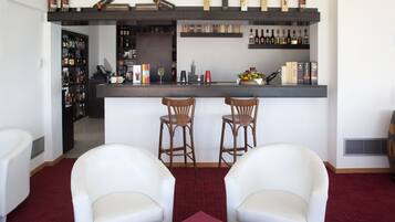 Una zona de bar con dos taburetes de madera, una barra con varias botellas y un frutero, y un espacio de estar con dos sillas blancas.