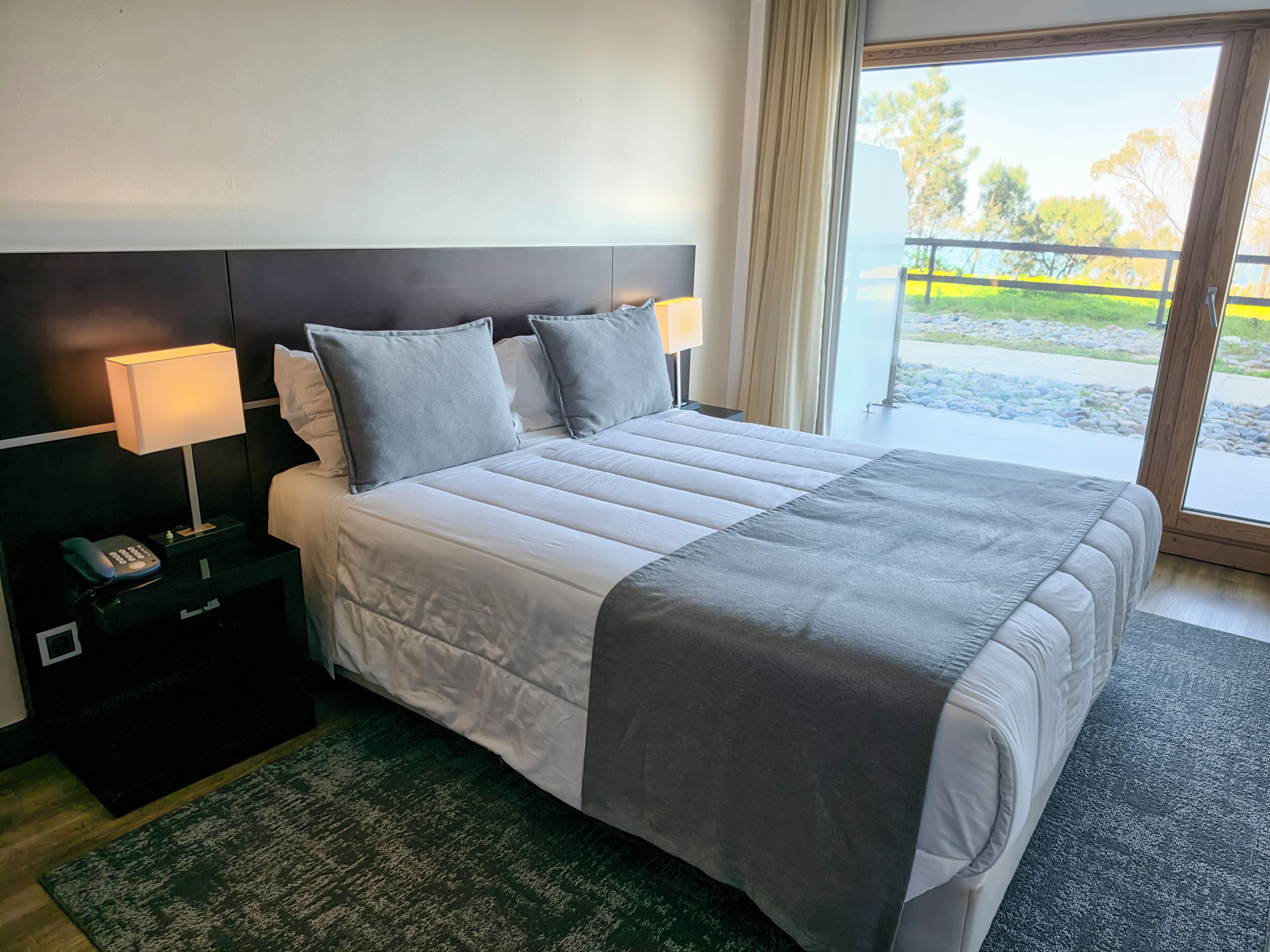 Habitación de hotel con una cama grande, lámparas de noche, un teléfono en la mesita y vistas al exterior.