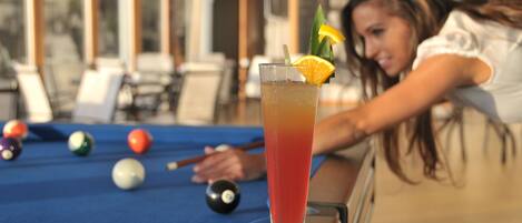 A pool table with a glass of iced drink garnished with an orange slice and a lemon twist, and a person leaning on the table.