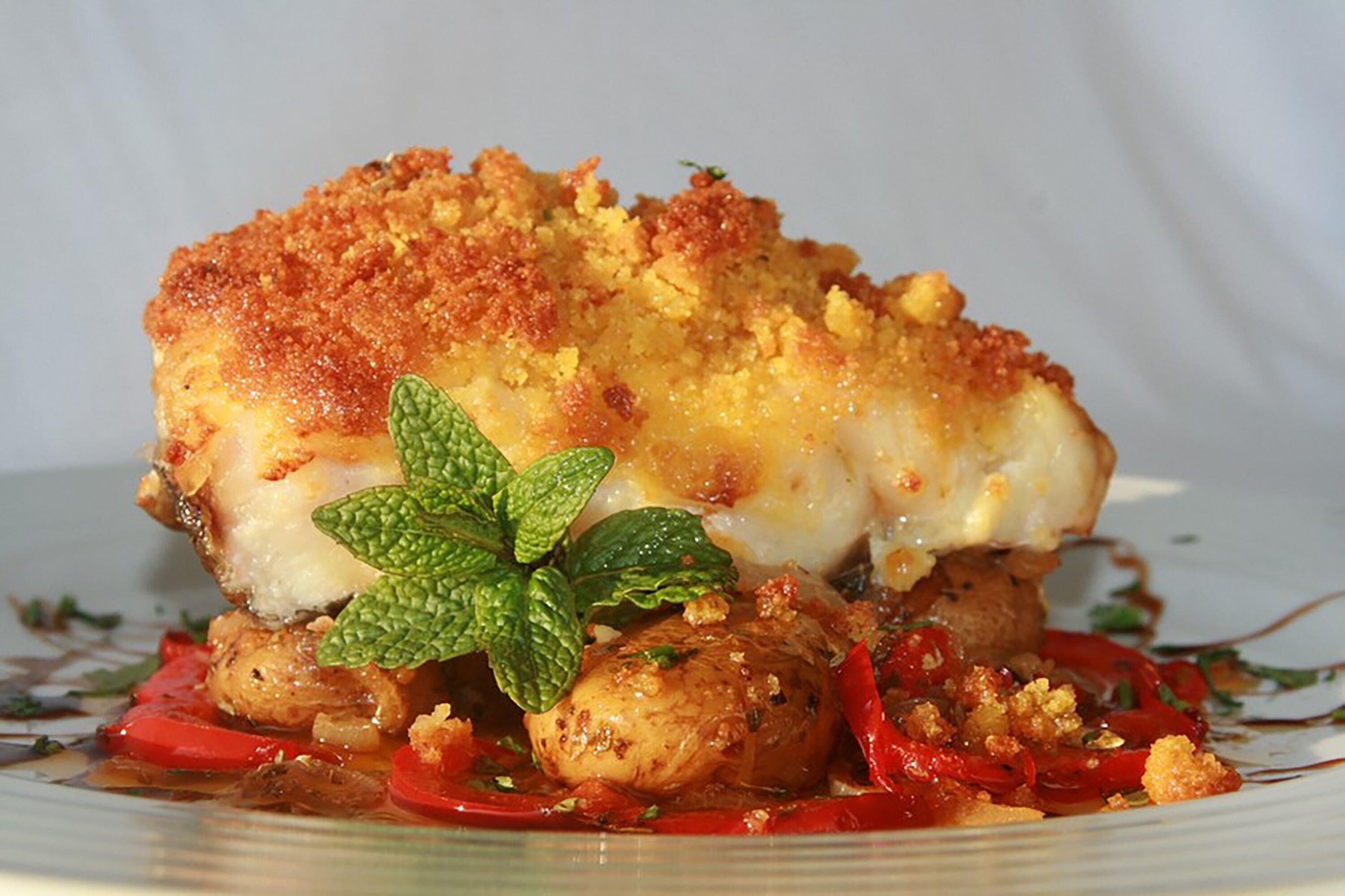 Un plato con costra dorada, salsa blanca y pimientos rojos, adornado con una hoja de menta.