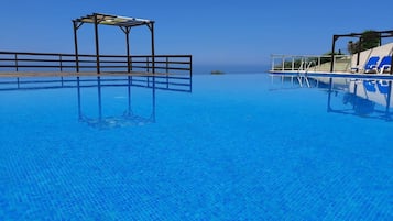 A clear blue infinity pool with a wooden deck and lounge chairs, overlooking a calm sea.