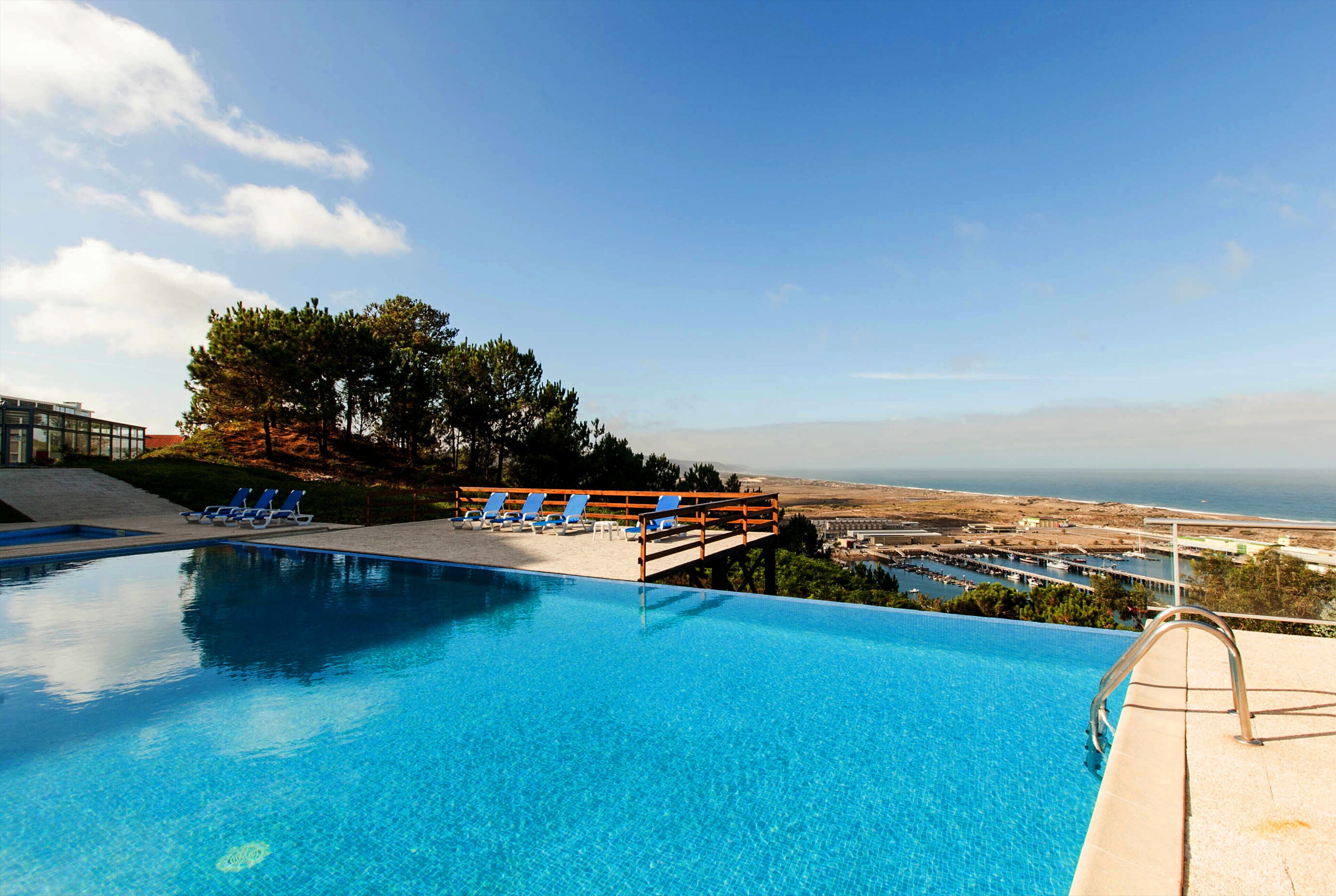 A clear blue pool with blue lounge chairs and a wooden deck overlooking a coastal view.