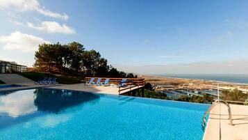 A clear blue pool with blue lounge chairs and a wooden deck overlooking a coastal view.