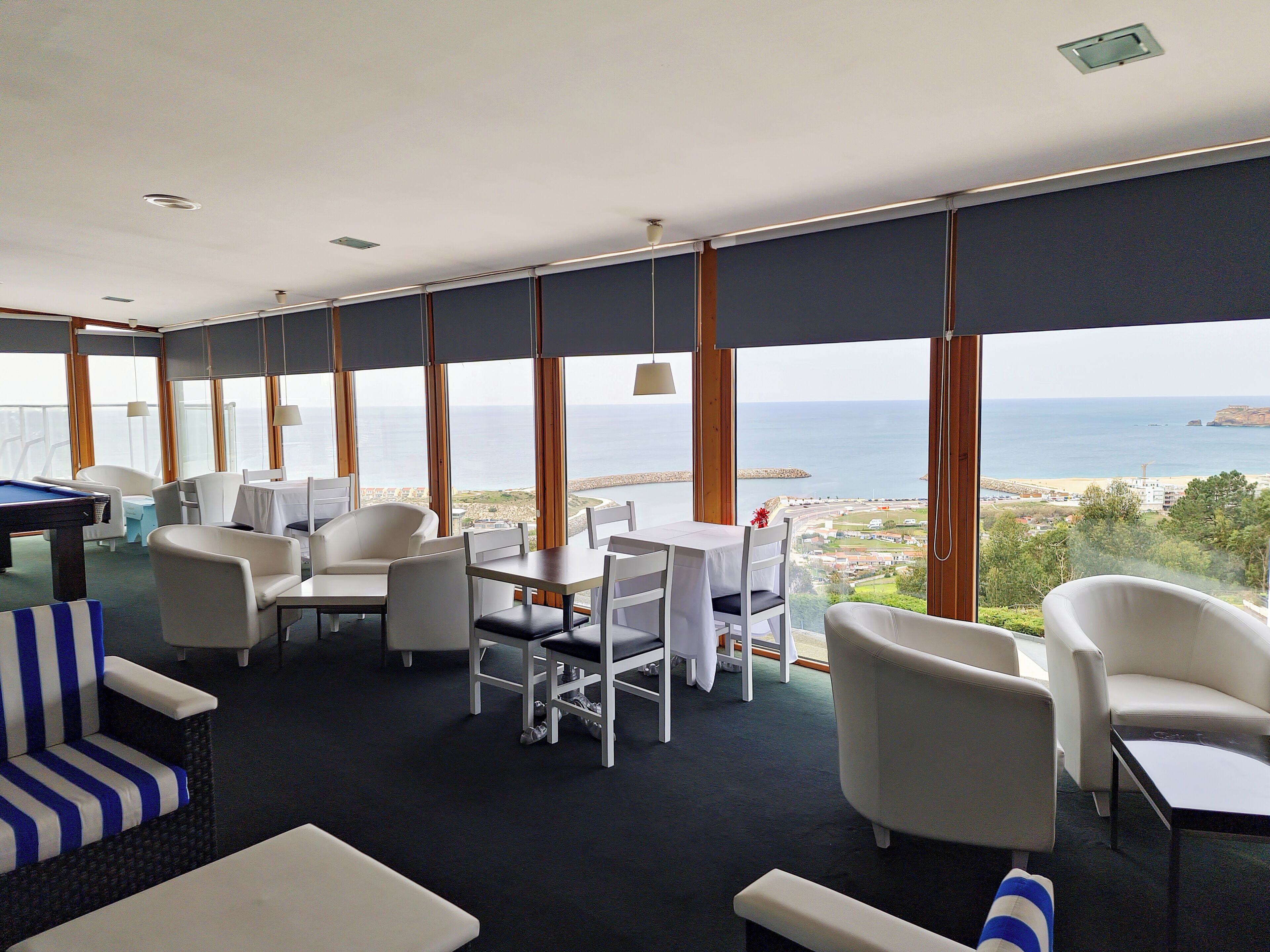 Une pièce comprenant une table de billard, des chaises blanches, une table à manger et de grandes fenêtres offrant une vue sur la mer et une ville côtière.