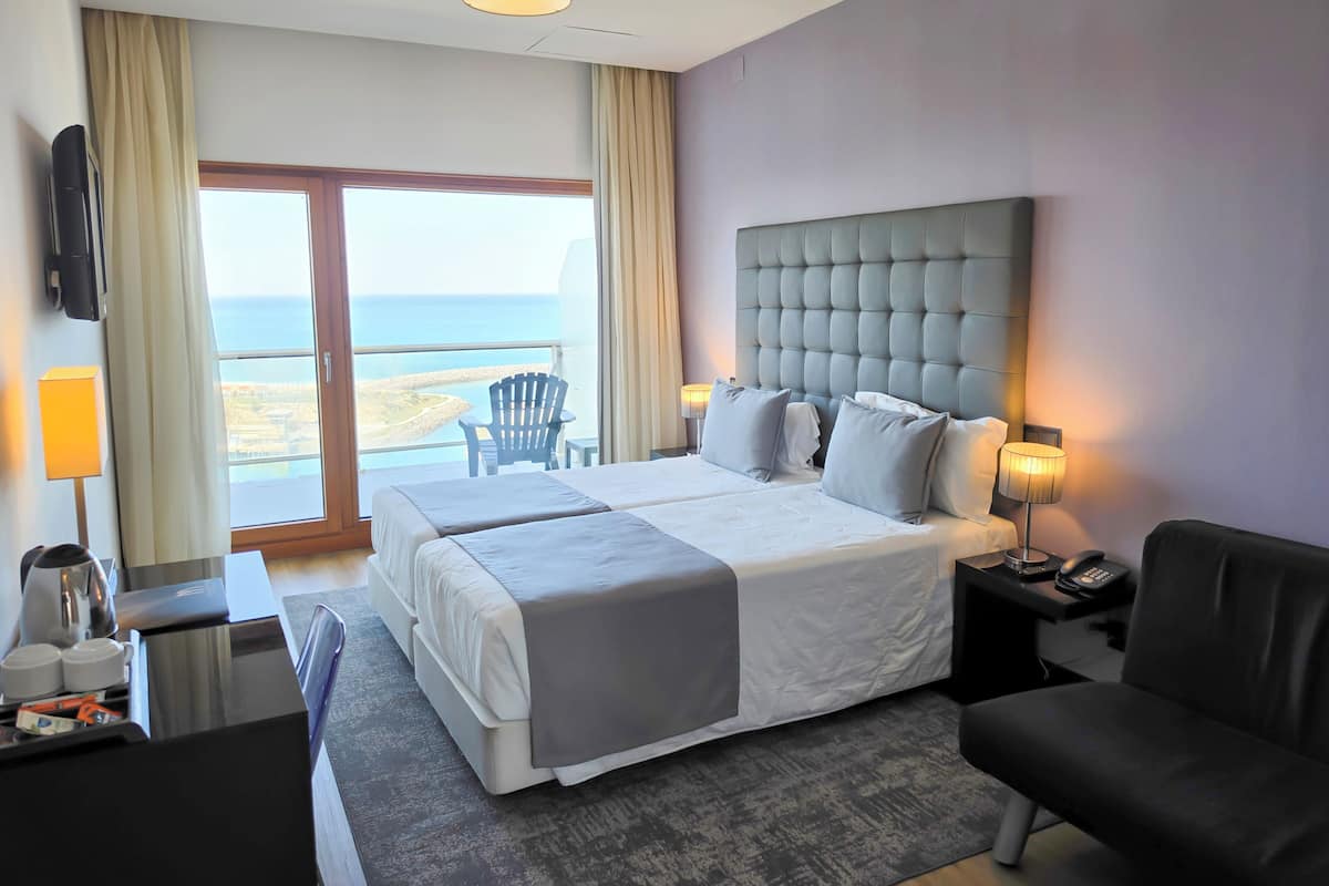 A hotel room with a large bed, a chair, a desk, a lamp, and a view of the sea.